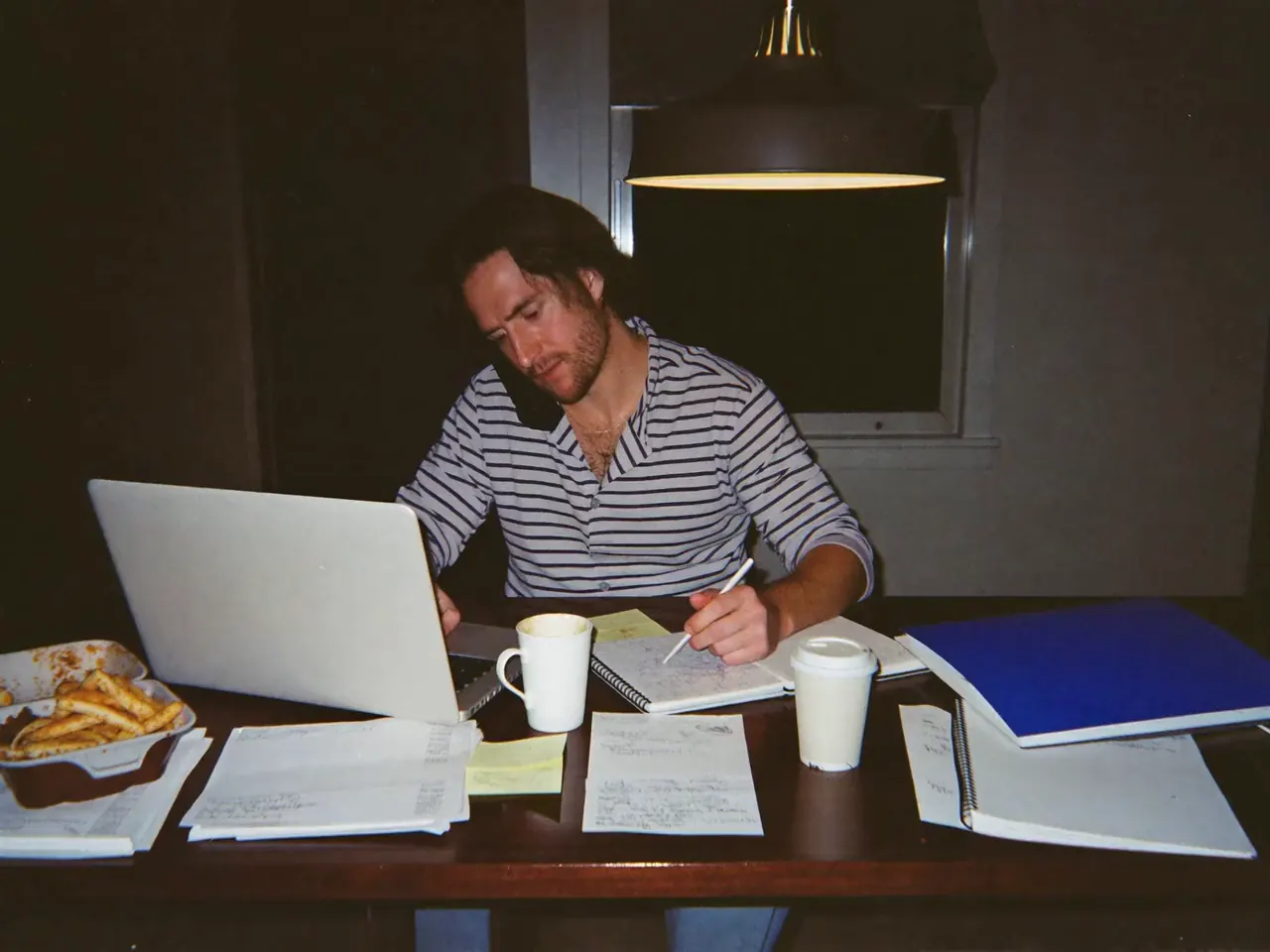Founder still working at his kitchen table at 11pm, phone on his shoulder, laptop and paperwork everywhere